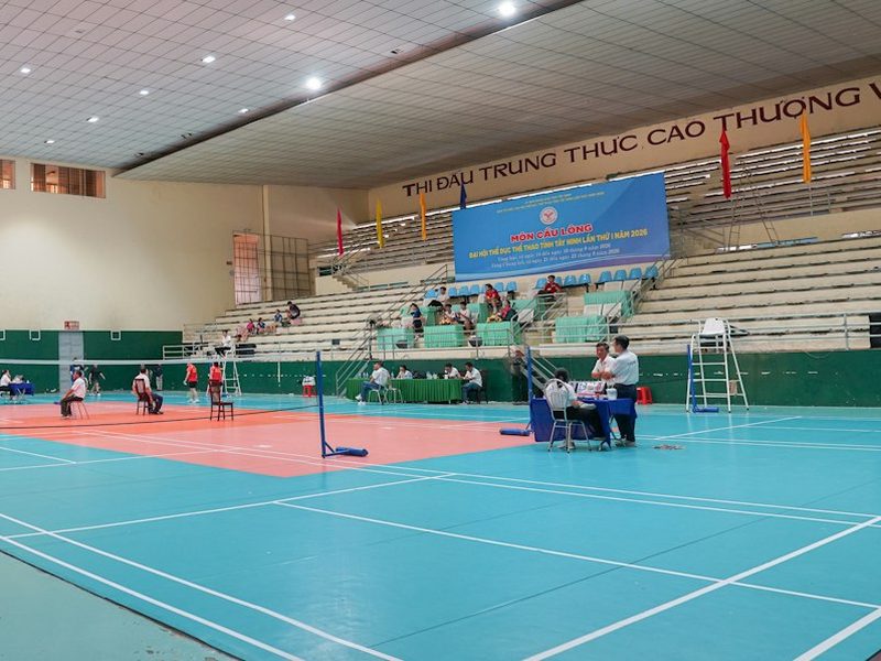 People on a badminton court inside a large indoor sports arena with bleachers