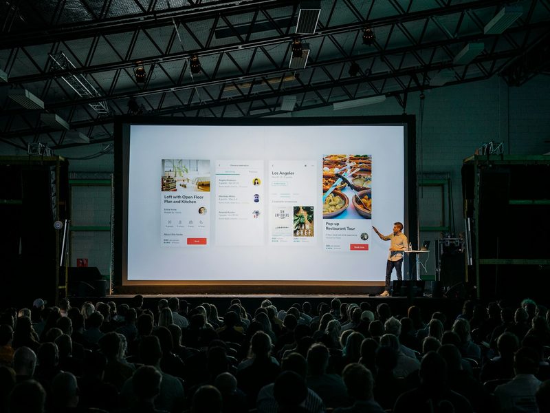 Person discussing while standing in front of a large screen in front of people inside dim- (illustrative photo)